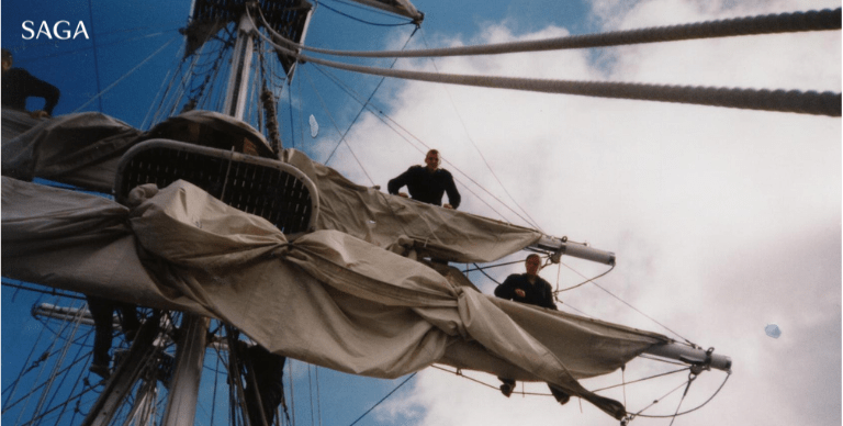 Captain Steve and other sit amongst a ships rigging. The sky is blue behind them.